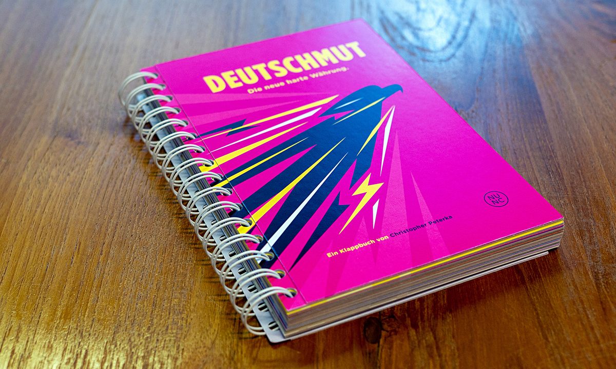 Standard-Ausgabe DEUTSCHMUT: magentafarbenes Klappbuch mit gelbem Titel auf Holztisch